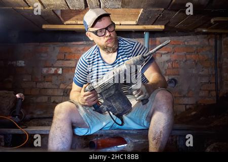 Lustiger bärtiger Mann in einem gestreiften T-Shirt und einer Sicherheitsbrille mit robuster Perforation hat Spaß bei Renovierungsarbeiten im Keller des Hauses. Hoch Stockfoto