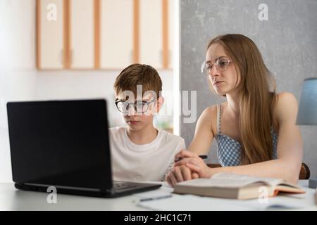 Das Kind ist online mit seiner Mutter beschäftigt und macht Hausaufgaben am Computer. Der junge Tutor arbeitet mit einem Studenten zusammen. Zu Hause Schulbildung. Stockfoto