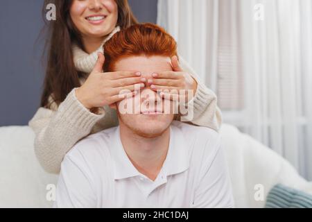 Der junge Mann, der auf dem Sofa saß, und seine Freundin, die hinter ihm stand, schloss seine Augen mit lächelnden Handflächen. Stockfoto