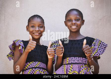 Zwei schöne schwarze afrikanische Schwestern in Zwillingskleidung, die mit Daumen nach oben auf die Kamera schauen; Girl Power Concept Stockfoto