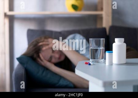 Frau liegt auf der Couch und leidet an Migräne. Flaschen mit Pillen und Glas Wasser gegen Mädchen, die an Kopfschmerzen leiden Stockfoto