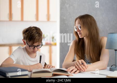 Junge mit Brille ist hart bei der Arbeit, seine Hausaufgaben zu schreiben. Ein junger Tutor hilft dem Schüler beim Unterricht. Zu Hause Schulbildung. Fernschulung Stockfoto