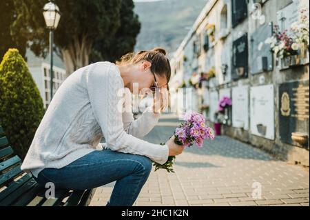 Traurige weinende Frau, die auf einer Bank mit Blumen auf dem Friedhof sitzt Stockfoto