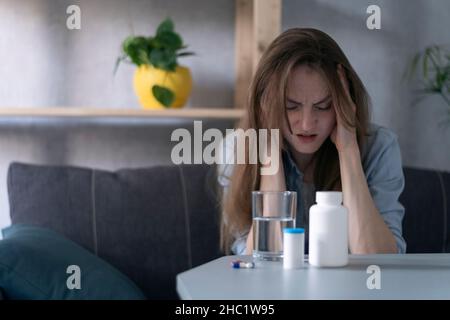 Frau sitzt auf der Couch und leidet an Migräne. Flaschen mit Pillen und Glas Wasser von Kopfschmerzen. Selbstmedikation zu Hause Stockfoto