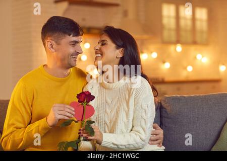 Der Ehemann umarmt seine geliebte Frau und gibt ihr eine Rose und eine Valentinskarte, während er auf der Couch sitzt. Stockfoto