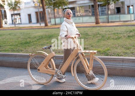 Vollkörper blonde Frau in Oberbekleidung schieben Holz Öko Fahrrad beim Gehen auf dem Bürgersteig in der Nähe von Rasen im Stadtpark. Stockfoto