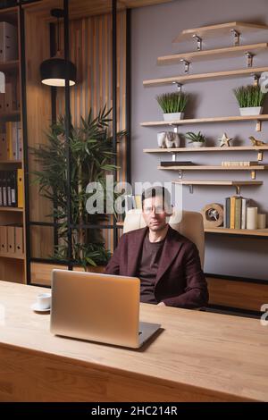 Ein junger ernsthafter Mann in burgunderrotem Anzug sitzt in einem Büro an einem Tisch vor einem Laptop und schaut in die Kamera. Dunkle Haare, braune Augen, weiße Haut. I Stockfoto