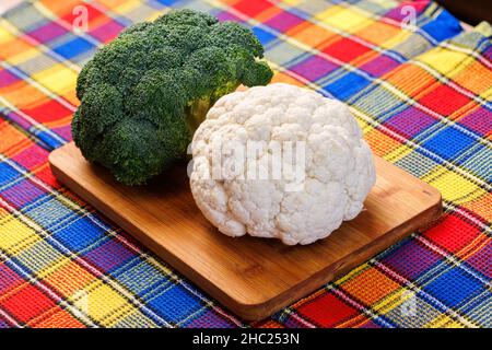 Ein Kopf aus Brokkoli und ein Kopf aus Blumenkohl liegen auf einem Schneidebrett auf dem Tisch, darunter ein buntes Handtuch Stockfoto