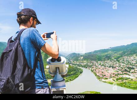 Männlicher Tourist in blauem Hemd steht am Fernglas des Aussichtspunkts, hält Handy und macht ein Foto von Mzcheta und dem Fluss Mgvari. Reisen Stockfoto