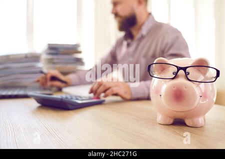 Nahaufnahme eines Sparschweins in Form eines rosafarbenen Schweins mit Brille, die auf dem Tisch auf dem Hintergrund eines geschäftigen Mannes steht. Stockfoto