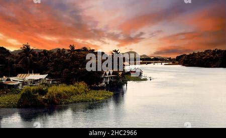 Dunkler Kontrastuntergang in den Tropen auf dem großen Fluss mit Dorf, Brücke und Bergen im Hintergrund. Die Brücke am Fluss Kwai, Thailand 5th. juli 2009. Hochwertige Fotos Stockfoto