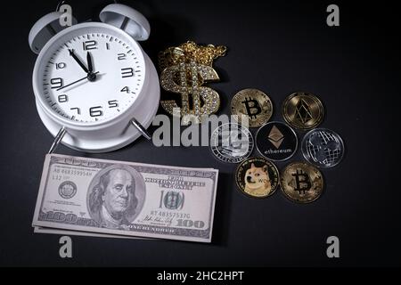 Einige verschiedene Krypto-Münzen liegen neben einem Stapel von 100-Dollar-Scheinen, einem weißen Wecker und einem diamantbesetzten Dollaranhänger Stockfoto