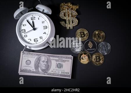 Einige verschiedene Krypto-Münzen liegen neben einem Stapel von 100-Dollar-Scheinen, einem weißen Wecker und einem diamantbesetzten Dollaranhänger Stockfoto