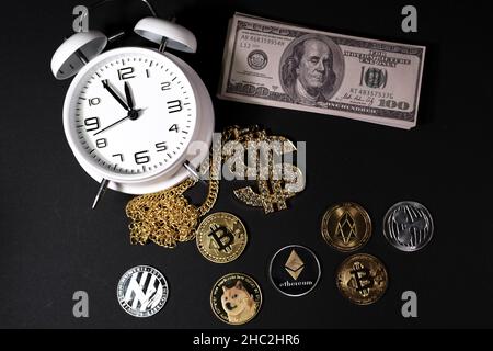 Einige verschiedene Krypto-Münzen liegen neben einem Stapel von 100-Dollar-Scheinen, einem weißen Wecker und einem diamantbesetzten Dollaranhänger Stockfoto