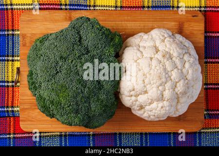 Ein Kopf aus Brokkoli und ein Kopf aus Blumenkohl liegen auf einem Schneidebrett auf dem Tisch, darunter ein buntes Handtuch Stockfoto