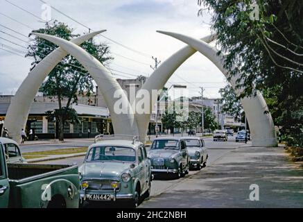 Die Mombasa Stoßzähne (oder Mapemba ya Ndovu oder Pembe za Ndovu ¬ die Suaheli für Elefantenzähne), ein Denkmal über der Moi Avenue, Mombasa, Kenia, abgebildet im Jahr 1967. Der Bogen wurde 1952 zu einem Gedenken an den Besuch der britischen Königsfamilie erbaut und bestand ursprünglich aus zwei hölzernen und leinwand-Stoßzähnen. Als die Straße zu zwei Fahrbahnen ausgebaut wurde, wurde 1956 vom stadtrat ein neuer Satz von vier witterungsbeständigen Aluminium-Stoßzähnen gebaut. Dann bildeten sie den Buchstaben ‘M’. Dieses Bild stammt von einem alten Amateur 35mm Kodak Farbtransparenz - ein Vintage 1960s Foto. Stockfoto