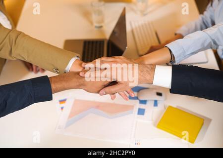 Junge Mitarbeiter sitzen am Schreibtisch in entspannter Atmosphäre beim morgendlichen Meeting im Büro. Menschen, die sich die Hände binden. Multirassisches Teamkonzept Stockfoto