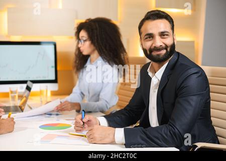 Portraitansicht des zufriedenen bärtigen multirassischen Geschäftsmannes, der zur Kamera lächelt, während eine ausscheidende Dame Papiere in der Hand hält und in ihrem Arbeitsraum arbeitet Stockfoto
