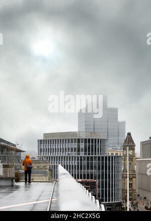 Anonymer Mann hoch oben in hellem Mantel stand allein auf dem Dach und blickte auf die Wolkenkratzer der Stadt, niedrige Wolken und stürmisches Nebelwetter. Stockfoto