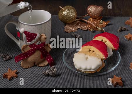 In eine Tasse wird heißes Wasser gegossen, um Tee zuzubereiten, mit bunten Weihnachtsmannskeksen und anderen Weihnachtsdekorationen in der Nähe Stockfoto