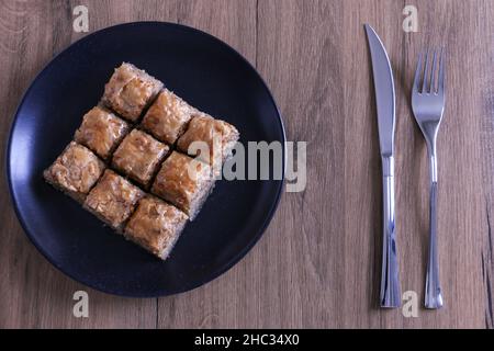 Baklava, ein traditionelles türkisches Dessert, das als Baklava auf einem schwarzen Teller auf einem hölzernen Hintergrund benannt wurde. Selektiver Fokus auf die zentrale Baklava. Ramadan-Konzept Stockfoto