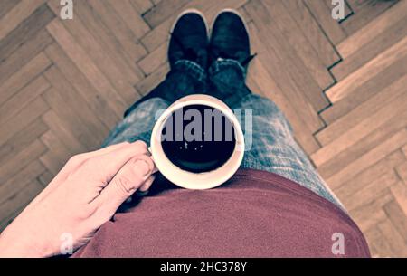 Nahaufnahme einer Hand, die eine Tasse Kaffee hält. Der Mann hält eine Tasse Kaffee am Morgen. Mann mit einer Tasse Kaffee auf dem Parkettboden. Nahaufnahme. Kopieren Stockfoto