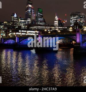 Die Lichter der Stadt spiegeln sich in der Nacht im Winter in der Themse mit einer Brücke und Wolkenkratzern im Hintergrund, London. Stockfoto