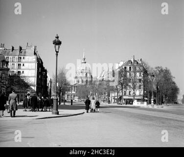 Paris Les Invalides von der Metro Saint-Francois-Xavier, 1945. Ein friedliches Bild, in dem viele Menschen, vielleicht nach der Kirche, unterwegs sind. Fast keine Fahrzeuge sind in Sicht und eine Familie mit einem Kinderwagen ist direkt auf der Straße zu Fuß dargestellt. Die Kamera befindet sich gegenüber dem Place Andre-Tardieu und blickt direkt auf die Avenue de Villars auf die große Barockkuppel. Der Blick geht weiter nach Norden auf den Boulevard des Invalides auf der rechten Seite. Bäume sind im frühen Frühjahr Laub, so dass es klare Sichtlinien. Stockfoto