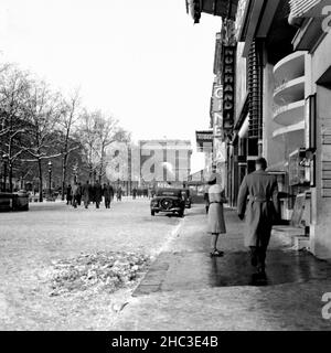 Paris Avenue des Champs-Elysees an der Metro George V mit Blick auf den Arc de Triomphe im Winter 1945. Eines von zwei Fotos, die Menschen beim Gehen und die Geschäfte rund um die U-Bahn mit Schnee auf dem Boden zeigen. Es sind fast keine Fahrzeuge in Sicht, und die Fotos wurden aufgenommen, als die Champs-Elysees noch mit baumgesäumten Medianen ausgestattet waren, die den Boulevard in eine bessere Fußgängerwaage einengen. Viele der Menschen sind in Militäruniform. Auf der nordöstlichen Seite der Avenue sind unter anderem das Café George V, die Normandie, Milady, Lord Byron und das Kino zu sehen. Die Bäume sind mit Schnee beladen. Stockfoto