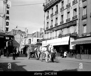 Mann, der auf einer Pariser Straße ein Paar weißer Dray-Pferde und einen Bauernwagen führt und entlang geht, 1945. Die Wagenräder sind fast so groß wie der Mann selbst. Um diesen Mann herum können etwa zehn weitere Pariser auf dieser nicht identifizierten Pariser Straße sehen, wie sie ihre Geschäfte machen. Das einzige Fahrzeug befindet sich direkt hinter dem Wagen. Kommerzielle Zeichen lesen Damoy, P. Bourgeon, Fleurist, Jean Morin; Produits alimentaires, vins Geldstrafen. Stockfoto