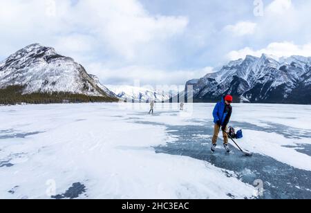 Junge, der im kalten Winter Eishockey auf dem gefrorenen Lake Minnewanka in den kanadischen Rockies des Banff National Park übt, mit nicht identifizierbaren Besuchern Stockfoto