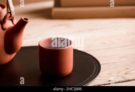 Tasse Tee oder Kaffee mit heißem Dampf aus braunem Ton, auf Holztisch mit teilweiser Ansicht der Teekannen oder Kaffeekannen. Bücher. Stockfoto