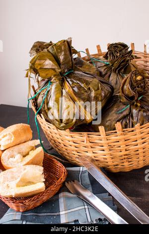 Traditionelles kolumbianisches Essen, bekannt als „Tamal“, verpackt mit Blättern in einem Korb auf einem schwarzen Tisch Stockfoto