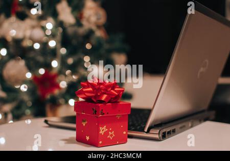 Kleines rotes Geschenk mit Geschenkschleife neben dem Laptop auf dem Tisch vor dem geschmückten weihnachtsbaum Stockfoto