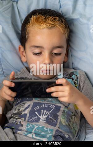 Eingetaucht kleinen Jungen in Freizeitkleidung liegend auf dem Bett und spielen Videospiel auf dem Handy, während Freizeit zu Hause verbringen Stockfoto