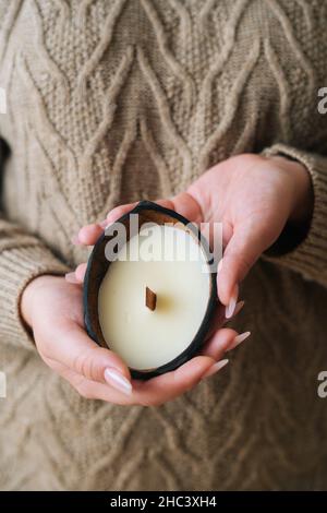 Vertikale Nahaufnahme einer nicht erkennbaren jungen Frau, die handgemachte Duftkerze in einer natürlichen schwarzen Schale in der Nähe der Brust hält. Stockfoto