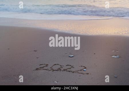 2022 Happy New Year Inschrift Sandstrand. Der geschriebene Text ist auf gelbem Sand geschrieben und wird von einer Welle weggespült. Das Konzept der Veränderung des Jahres, Stockfoto