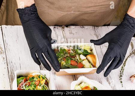 Hände in schwarzen Handschuhen packen ein Business Lunch in einen Karton. Konzept Food Delivery Service. Leckere und gesunde Lebensmittel am Arbeitsplatz. Tak Stockfoto