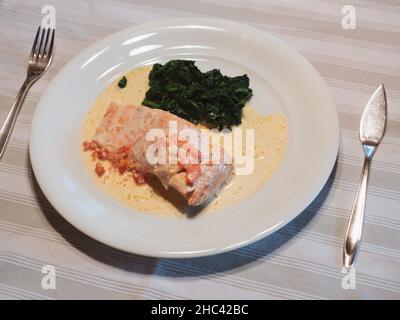 Lachs in Sahnesauce mit gebuttertem Spinat auf einem weißen Teller mit Silberfischmesser und Gabel Stockfoto