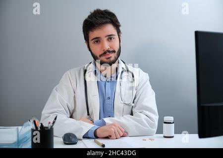 Junger Arzt, der die Kamera anschaut und lächelnd am Schreibtisch in einem medizinischen Büro sitzt. Stockfoto
