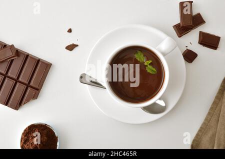 Heiße Schokolade in weißem Keramikbecher, Süßwarenschokolade und Stücke und Pulver in Schüssel auf weißem Tisch. Draufsicht. Horizontale Zusammensetzung. Stockfoto