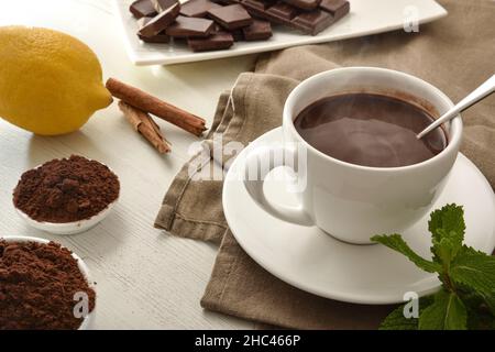 Heiße Schokolade in weißem Keramikbecher, Stücke und Schokoladenpulver in Schale auf Holztisch. Erhöhte Ansicht. Horizontale Zusammensetzung. Stockfoto