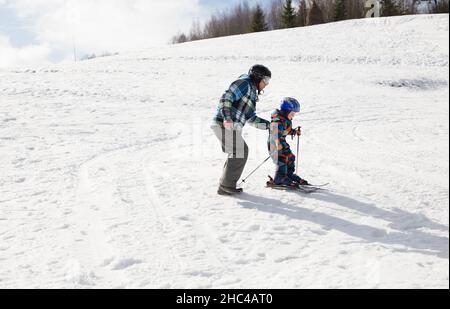 Kind mit Vater lernt während des Winterurlaubs in verschneiten Bergen an sonnigen, kalten Tagen Skifahren. Winter aktiv Spaziergänge mit Kindern. Saisonale Freuden, fröhliches Chil Stockfoto