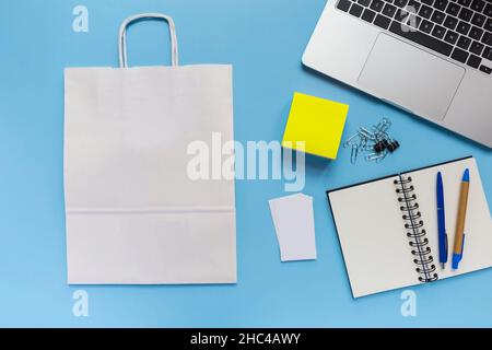 Flaches Lay, weiße Papiertasche, Arbeitsbereich mit blauem Hintergrund und Laptop, Notizblock, Visitenkarte und zwei Stifte, Schreibtisch Stockfoto