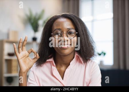 Lächelnde afroamerikanische Frau mit Brille und pinkem Hemd, die mit Büchern und Notizen am Schreibtisch sitzt und alles in Ordnung macht. Weibliche Tutorin, die während des Fernlernens zu Hause arbeitet. Stockfoto