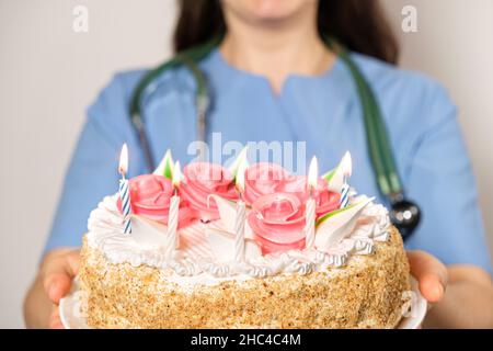 Eine Ärztin in blauer Uniform hält einen Geburtstagskuchen mit Kerzen und lächelt. Stockfoto