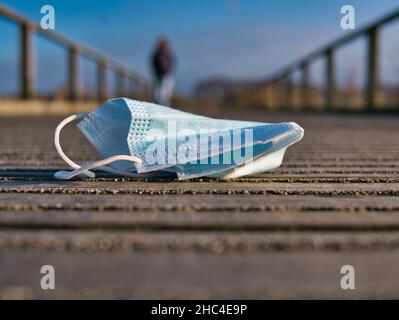 Eine blaue, weggeworfene Plastikmaske auf einer Holzpromenade in ländlicher Umgebung, wenn eine einzelne nicht identifizierbare Person weggeht. Stockfoto