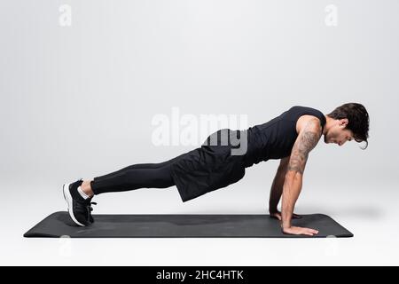 Seitenansicht des Sportlers in Rückensportbekleidung, der auf der Fitnessmatte auf grauem Hintergrund eine Planke macht Stockfoto