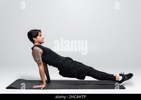 Seitenansicht des Mannes, der auf der Fitnessmatte auf grauem Hintergrund eine Planke macht Stockfoto
