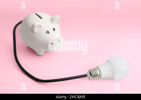 Weißes Keramik-Sparschwein mit Elektrodraht als Schwanz, verbunden mit Glühbirne, auf rosa Hintergrund. Konzept für Energiepreise, Inflation Stockfoto